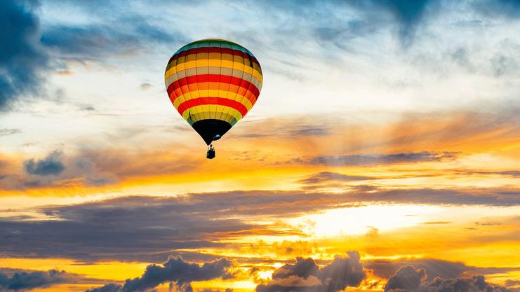 Bunter Heißluftballon schwebt über einem See bei Sonnenuntergang mit dramatischem Wolkenhimmel.