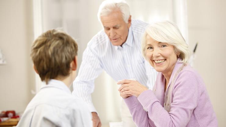 Lächelnde ältere Frau sitzt am Tisch, während ein Mann im Hintergrund Getränke serviert, ein Junge schaut sie an.